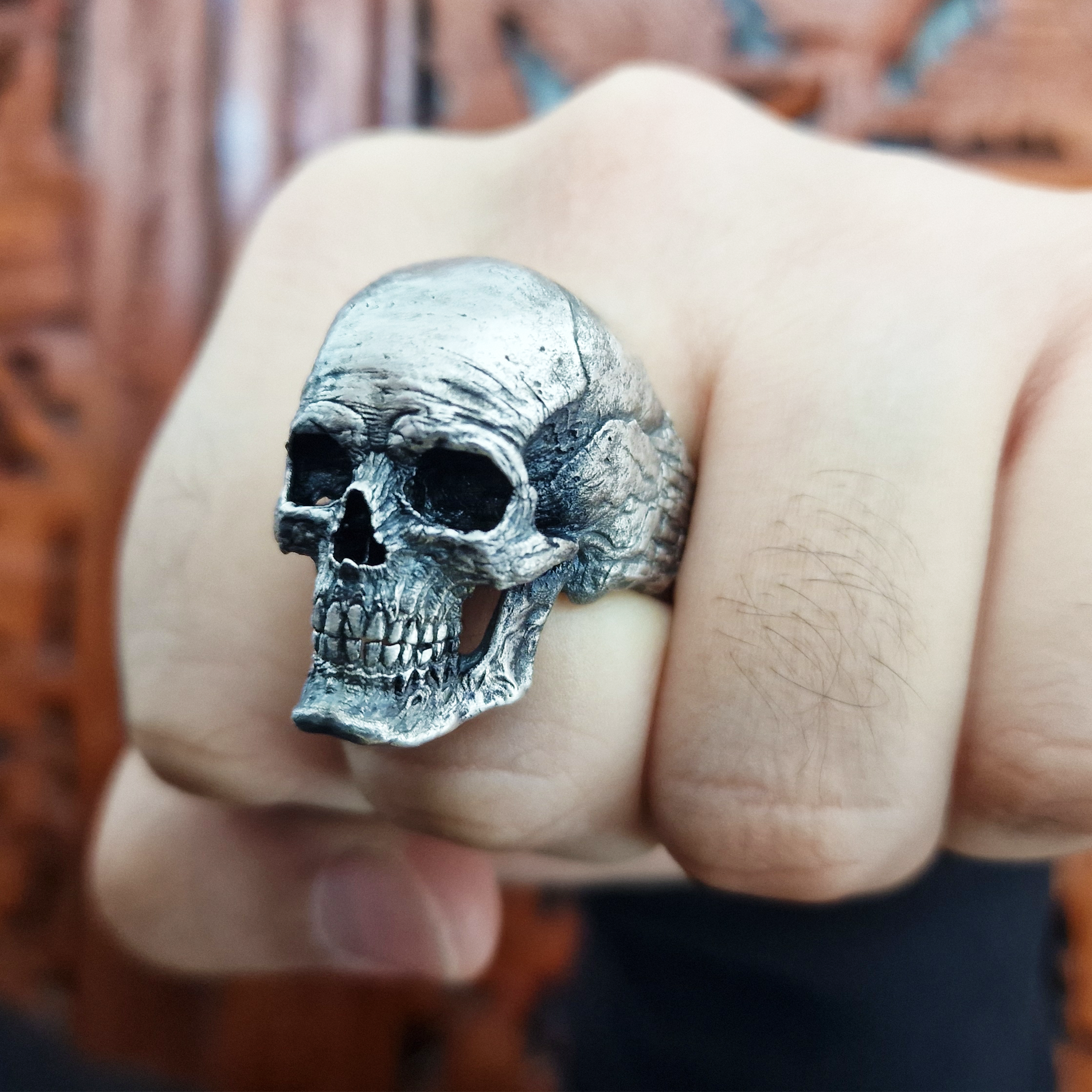 Fist pointed toward the camera in natural light, wearing the Invidia Mortis ring in the Fallen finish, with rich patina and detailed skull design.