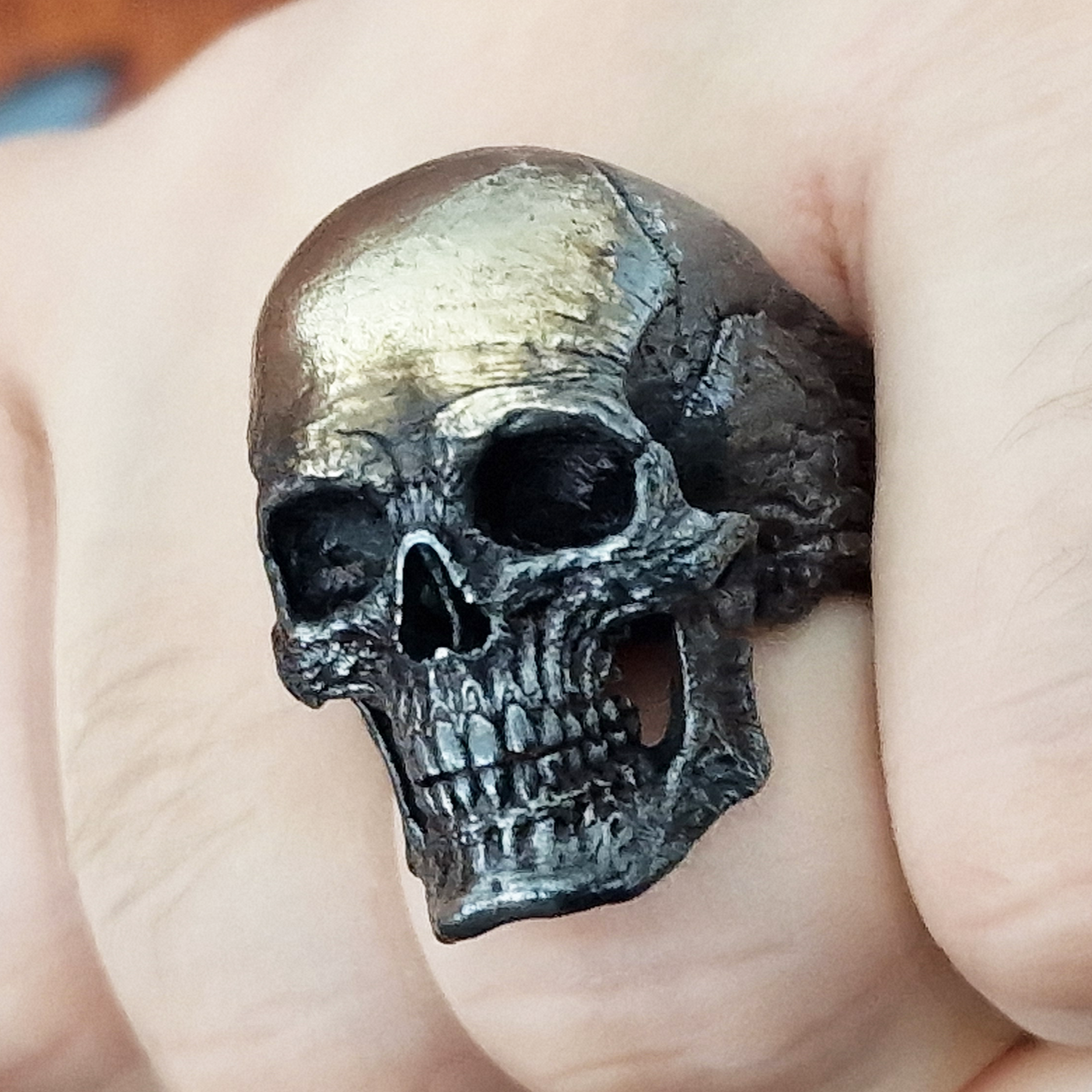 Fist pointed toward the camera in natural light, wearing the Invidia Mortis ring in the Revenant 1 finish, highlighting its details and texture.