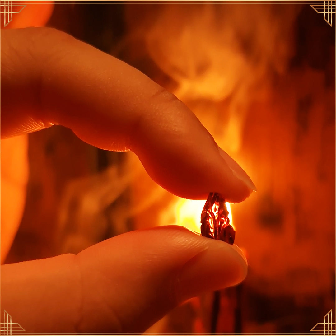 Close-up of a garnet gemstone held between pinched fingers, with candlelight and swirling smoke softly blurred in the background.